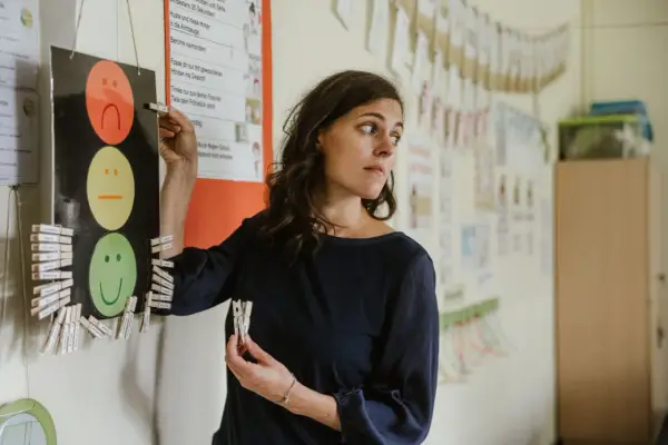 Lehrerin mit Klemmbrett und Namensschildern, die auf ein Ampelsystem mit Smiley-Gesichtern zeigen. Möglicherweise ein Hinweis auf Probleme mit der Schulleitung und Lehrern.